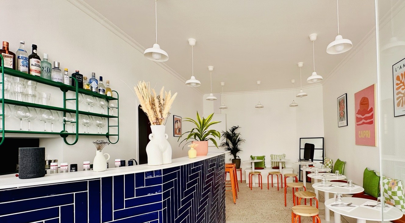 La salle ouverte au public : comptoir zellige bleu nuit, étagère verte garnie, tables blanches et tabourets orange.