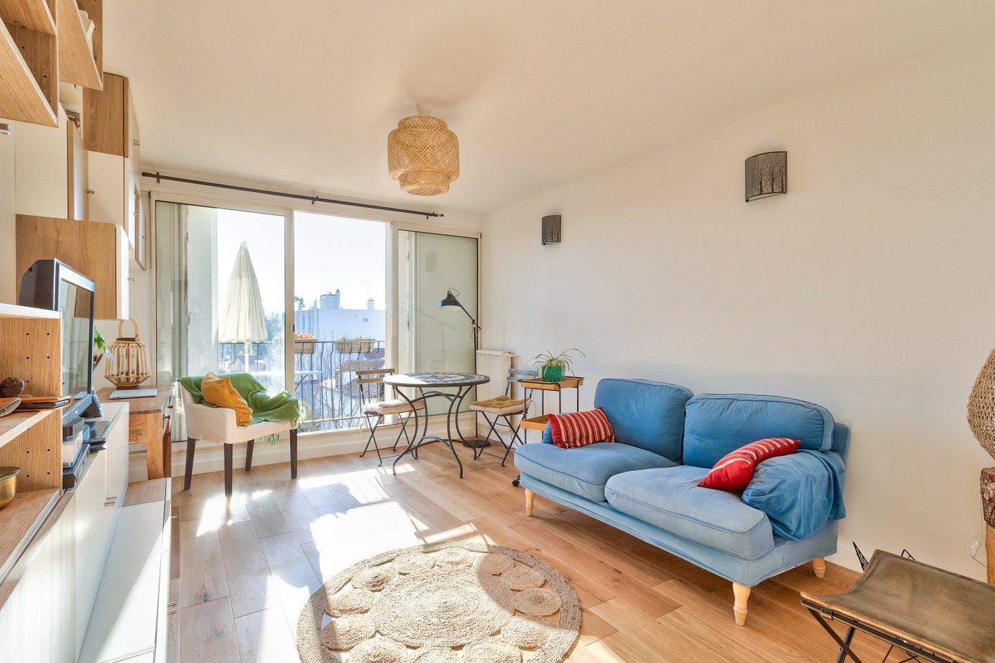 Séjour lumineux au parquet de chêne clair : canapé bleu, tapis de jute rond, suspension en rotin et baie vitrée ouvrant sur le balcon.