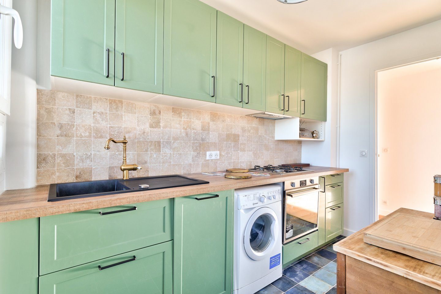 Cuisine vert sauge à crédence de travertin en damier, plan de travail en bois et robinetterie en laiton, sol en carreaux anciens bleu-vert.