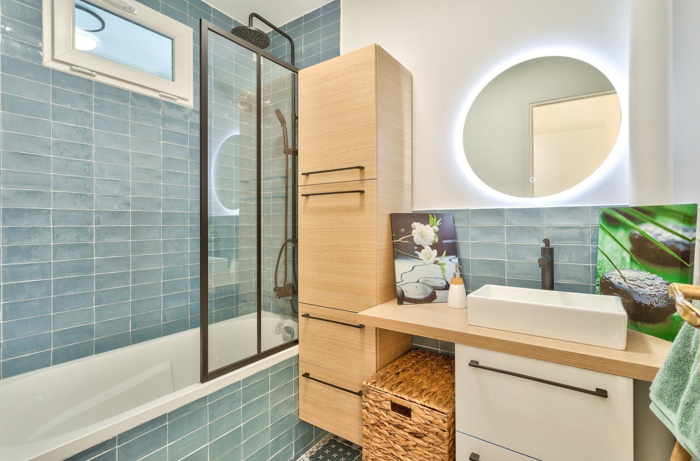 Salle de bain habillée de zellige bleu-vert : baignoire, colonne en chêne, miroir rond rétroéclairé et robinetterie noire.