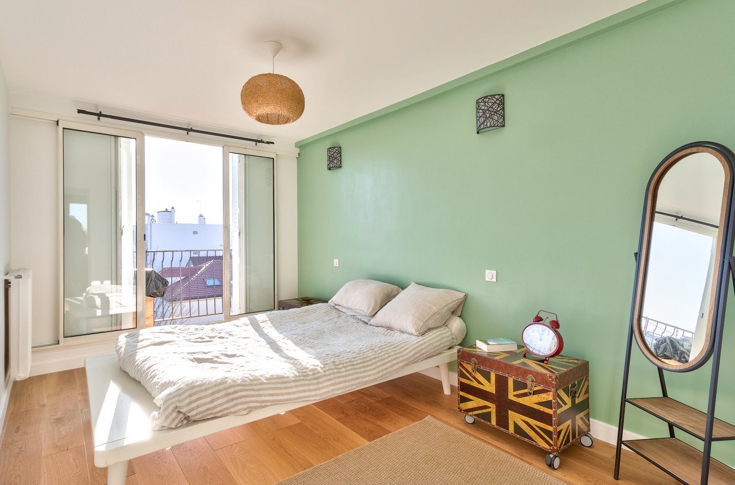 Chambre au mur vert d'eau, lit bas en linge rayé, miroir psyché et suspension en rotin, ouverte sur le balcon.